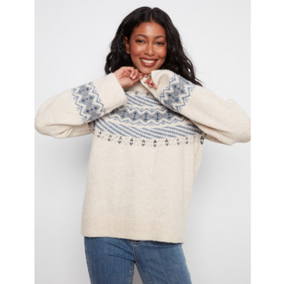 A woman wearing a cream-colored knit sweater with a blue and gray geometric pattern across the chest and cuffs, paired with blue jeans, against a light gray background.