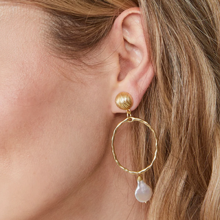 A close-up of a woman's ear wearing a gold hoop dangle earring with a textured gold top post and a teardrop-shaped white pearl drop.