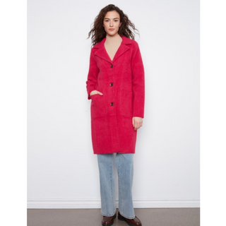 A woman modeling a bright pinkish-red mid-length coat with a soft textured finish, paired with light blue jeans and dark brown shoes, standing against a plain white wall.