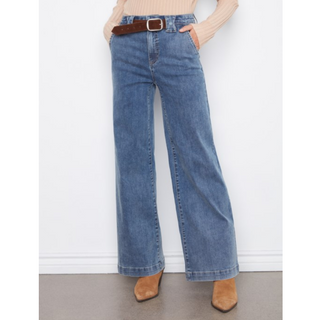 A person wearing blue wide-leg jeans with a brown belt, a beige ribbed top, and tan ankle boots, standing against a white wall.