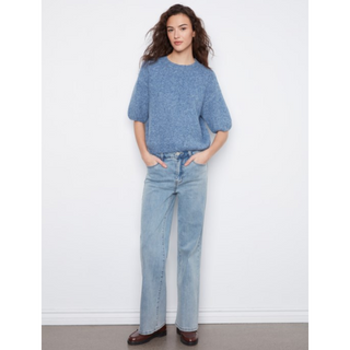A woman stands against a plain white wall wearing a light blue knit short-sleeve top and light blue straight-leg jeans, with brown shoes.