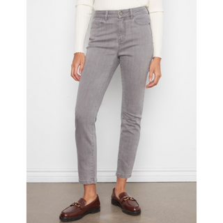 A person wearing light gray slim-fit jeans with a white long-sleeve top and dark brown leather loafers, standing against a white wall.