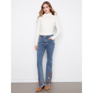 A woman modeling a white mock-neck long-sleeve knit top and blue flare jeans with colorful floral embroidery near the hem, paired with brown ankle boots, against a white wall.