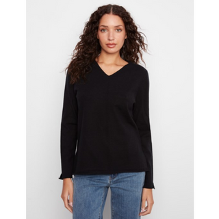 A woman with long, curly brown hair wearing a solid black long-sleeve V-neck top and blue jeans, standing against a light gray studio background.