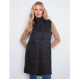A woman modeling a black sleeveless quilted puffer vest over a tan long-sleeve top and blue jeans, standing against a light gray studio background.
