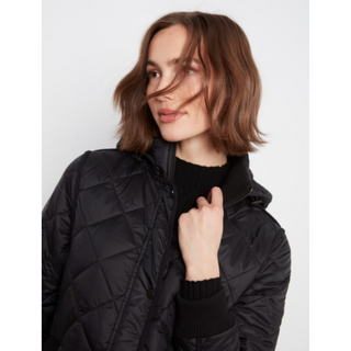 A woman with short brown hair wearing a black diamond-quilted puffer jacket over a black ribbed turtleneck sweater, photographed against a light gray studio background.
