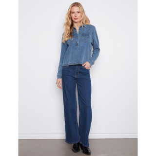 A woman modeling a light blue denim long-sleeve half-button shirt and dark blue wide-leg denim jeans against a plain white wall.