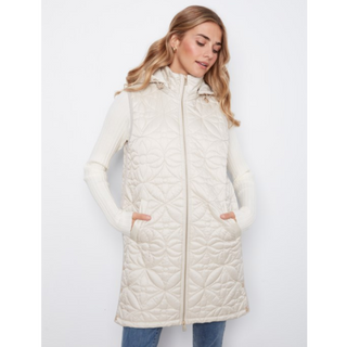 A woman wearing a long cream quilted puffer vest over a matching cream ribbed sweater and blue jeans, standing against a plain light gray studio background.