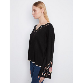 A woman shown in side profile wearing a black long-sleeve knit top with embroidered floral details on the lower sleeve and neckline, paired with blue jeans, against a plain light gray studio background.