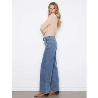 A woman wearing a beige ribbed knit sweater and wide-leg blue denim jeans, standing in profile against a plain white wall.