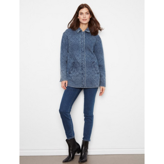 A woman modeling a blue denim jacket with an allover tonal floral or damask pattern, paired with dark blue skinny jeans and black ankle boots, standing against a plain white wall.