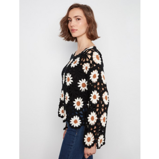 A woman wearing a black crochet top with a white and brown daisy floral pattern, paired with blue jeans, photographed against a light gray studio background.