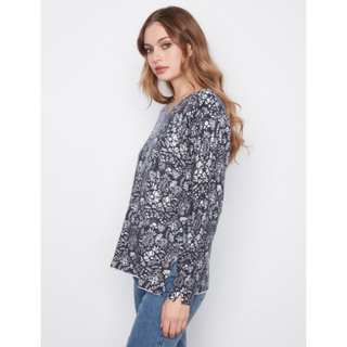 A woman wearing a long-sleeve, loose-fitting sweater with a black and white floral pattern, paired with blue jeans, photographed against a plain light gray background.