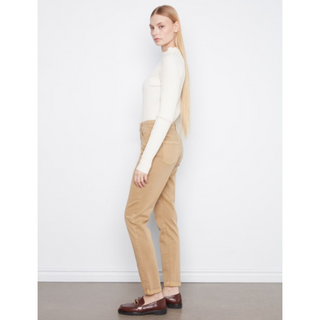 A woman stands in profile wearing a fitted cream long-sleeve top, tan straight-leg pants, and dark brown loafers against a plain white wall and gray floor.