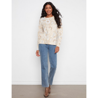 A woman modeling a cream-colored long-sleeve knit sweater with a beige floral print, paired with blue jeans and brown ankle boots, standing against a plain white wall.