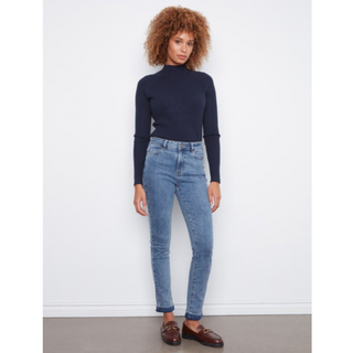 A model with curly brown hair stands against a white wall wearing a dark navy ribbed mock-neck long-sleeve top, light blue skinny jeans, and brown leather loafers.