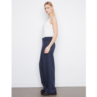 A woman stands in profile wearing a white sleeveless top and navy wide-leg trousers against a plain white wall.