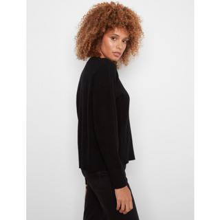 A woman with curly light brown hair is shown from a side-back angle wearing a black long-sleeve knit top and black pants against a plain light gray studio background.