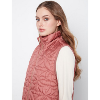 A woman wearing a sleeveless quilted puffer vest in dusty rose pink over a cream long-sleeve top, shown from the side against a light gray studio background.