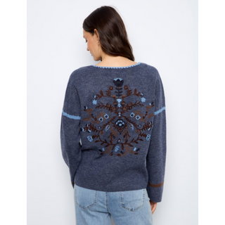 A woman viewed from the back wearing a dark slate blue knit sweater with a large brown and light blue floral pattern across the back, paired with light blue jeans, against a plain white background.