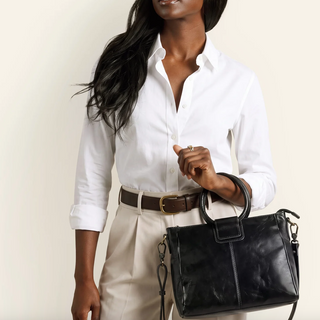 A woman wearing a white button-down shirt, beige trousers, and a brown belt carries a black leather handbag with top handles and a shoulder strap against a plain light background.